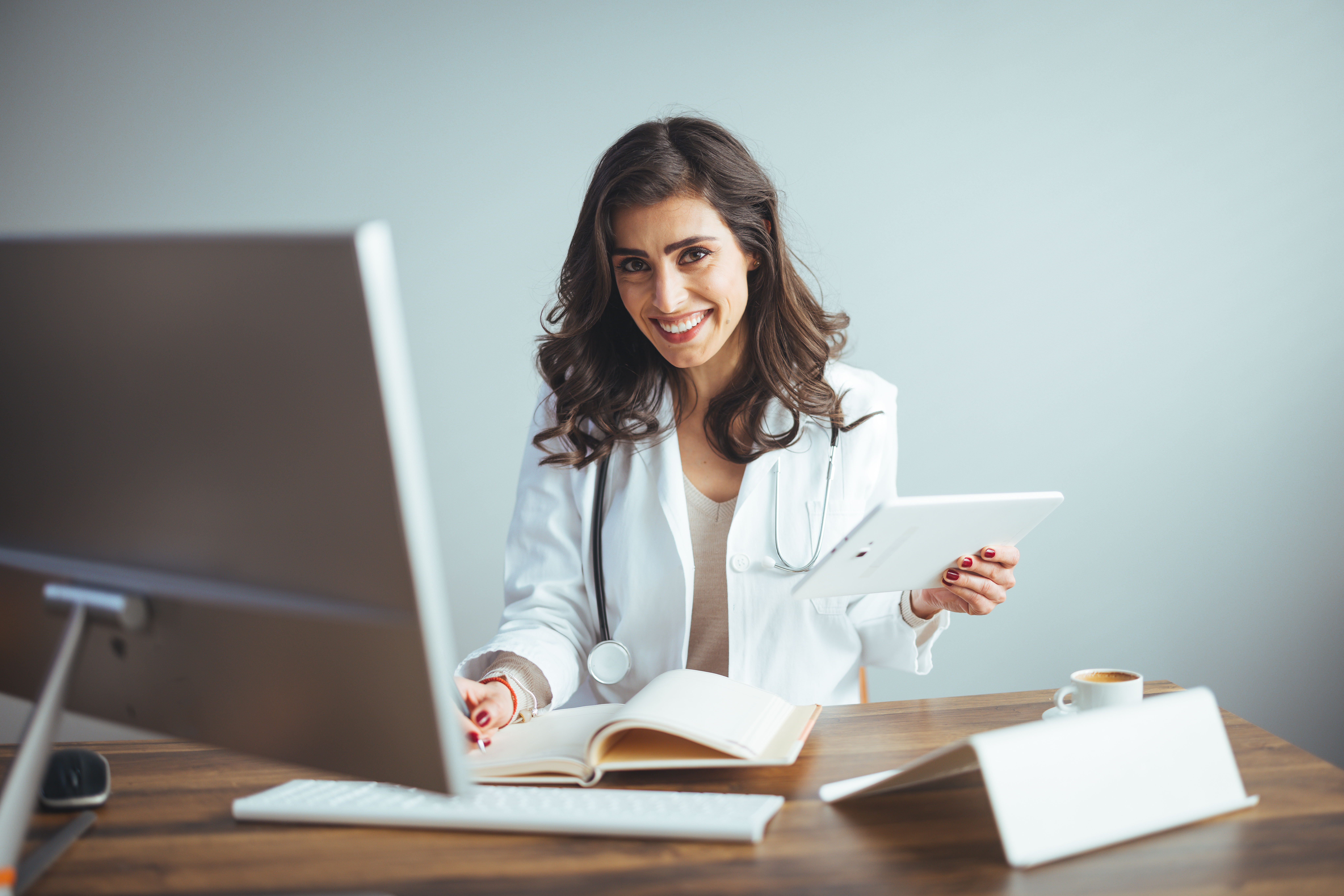 A woman sits at her desk in a labcoat and stethoscope!. She is smiling and looking past her computer towards the camera