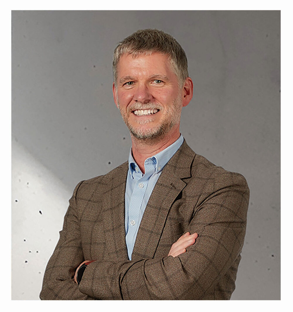 An image of Deanhallows in a brown jacket, he is smiling and has his arms crossed